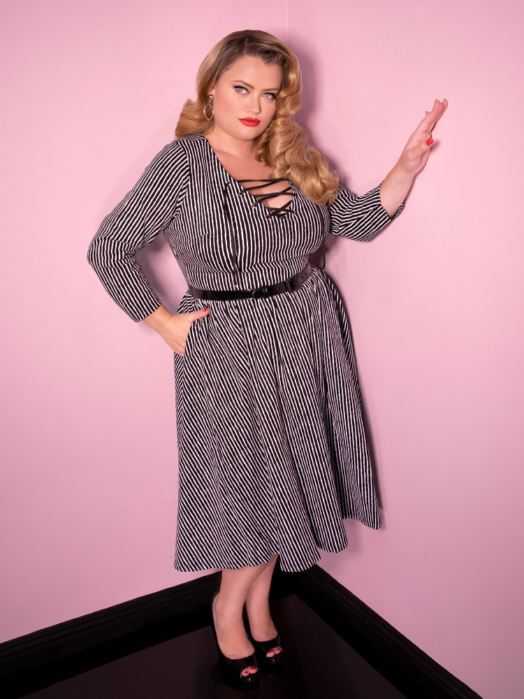 Blondie looking off camera with her hand in her pocket modeling the Vixen Clothing Miss Kitty swing dress in black stripes.