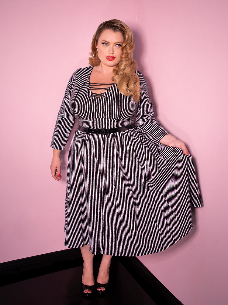 Blondie looking off camera and holding her skirt modeling the Vixen Clothing Miss Kitty swing dress in black stripes.