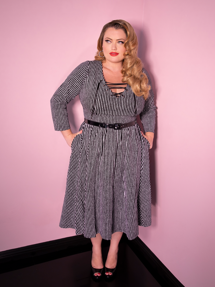 Blondie looking off camera with her hands in her pockets modeling the Vixen Clothing Miss Kitty swing dress in black stripes.